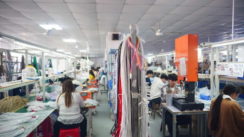 Inside a textile factory with workers seated at workstations handling materials. Bright fluorescent lighting fills the busy environment.
